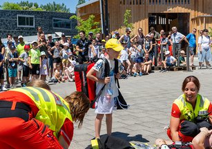 Ein Kind trägt den Crew-Bag und schaut der Rettungssanitäterin zu, wie sie die Figurantin versorgt.